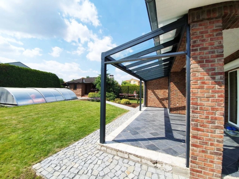 Moderní antracitová hliníková pergola se skleněnou střechou u cihlového domu se zahradou a bazénem.